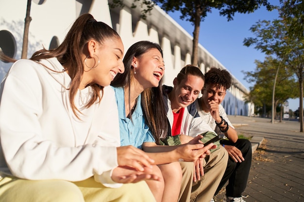 Группа счастливых друзей пользуется телефоном и веселится на улице. Молодые многорасовые люди смеются.