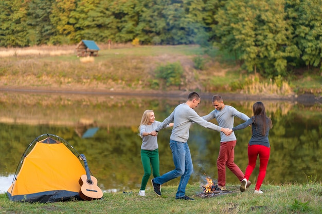 Group of happy friends with guitar, having fun outdoor, dancing and jumping near the lake in the park background the Beautiful sky. Camping fun