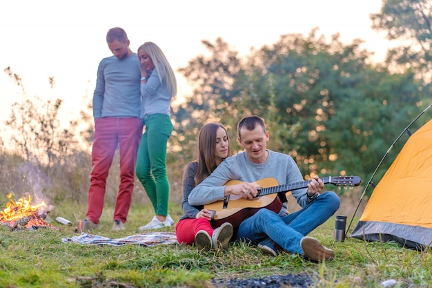 Group of happy friends with guitar, having fun outdoor, near bonfire and tourist tent. Camping fun happy family