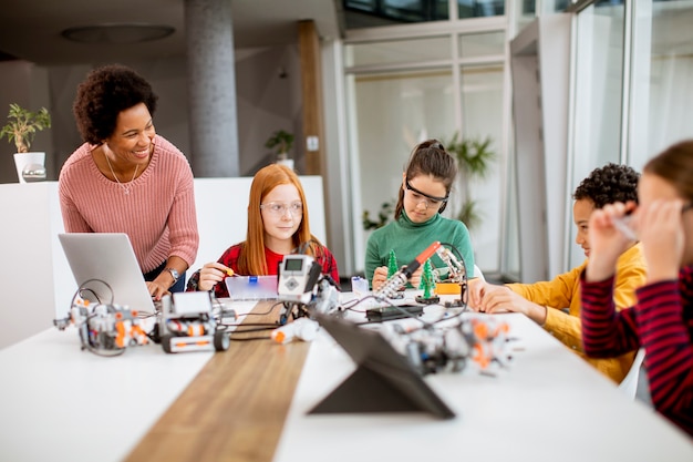 Группа счастливых детей со своей афро-американской учительницей естествознания с ноутбуком, программированием электрических игрушек и роботов в классе робототехники
