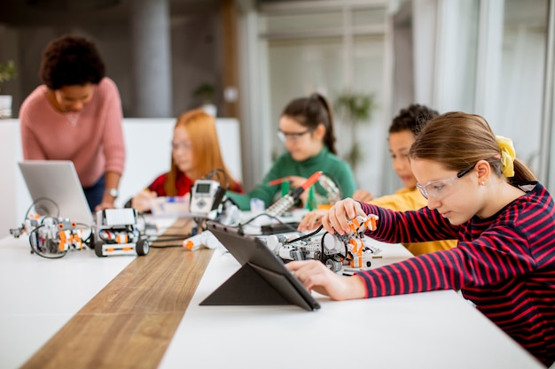 Gruppo di bambini felici con il loro insegnante di scienze femminile afroamericano con laptop che programmano giocattoli elettrici e robot in aula di robotica