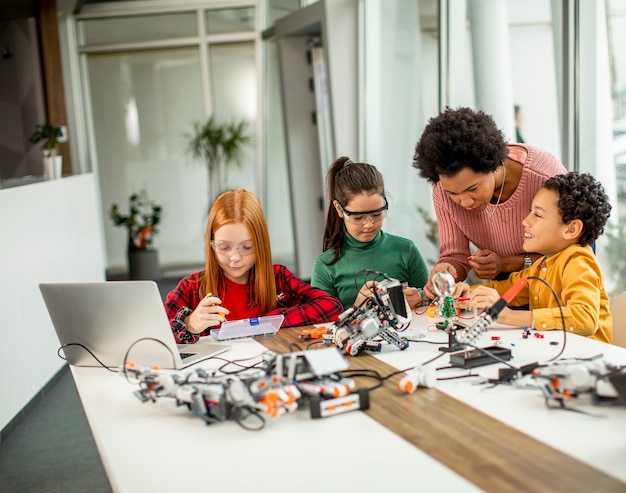 Gruppo di bambini felici con il loro insegnante di scienze femminile afroamericano con laptop che programmano giocattoli elettrici e robot in aula di robotica