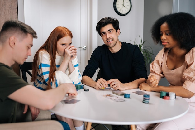 Photo group of happy multiethnic friends having home party on weekends playing poker game in living room sitting at table