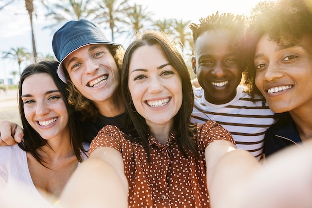 Foto gruppo di persone multirazziali felici che si fanno un selfie con il telefono cellulare all'aperto con la luce del sole posteriore