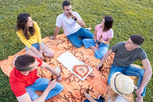 ピザを食べたり、瓶ビールを飲んだりする公園で幸せな人々のグループが、リラックスして社交する時間を持っているテイクアウトのミレニアル世代との即席の芝生パーティー