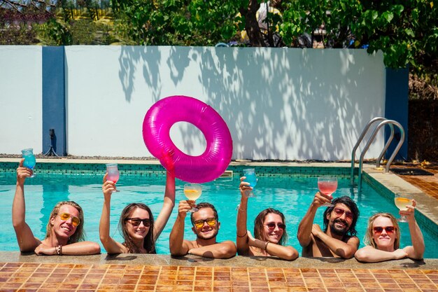 Group of happy six friends drinking jumping in pool day party with splash