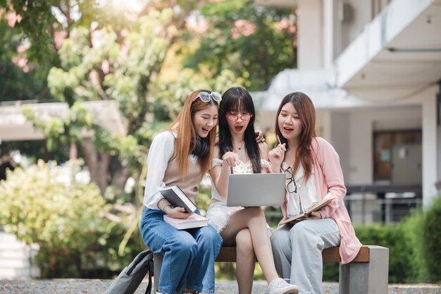Photo group of happy young asian college students sitting on a bench looking at a laptop screen discussing