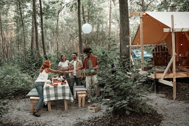 Group of happy young friends making barbecue in forest in glamping. African man grilling sausages passing camp food to his friend, laughing, having fun on a picnic in the countryside, grilling