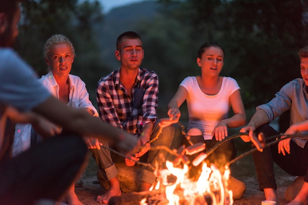 a group of happy young friends relaxing and enjoying  summer evening around campfire on the river bank