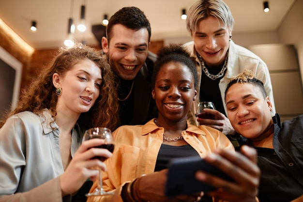 Group of happy young intercultural friends gathered in living room looking through new photos in sma