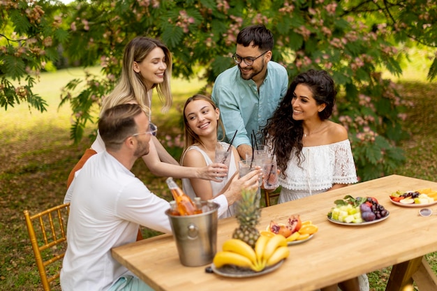 Gruppo di giovani felici che fanno il tifo con limonata fresca e mangiano frutta in giardino