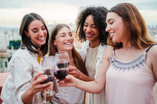 Photo group of happy young women friends having fun toasting wine glasses on a rooftop party drinking and