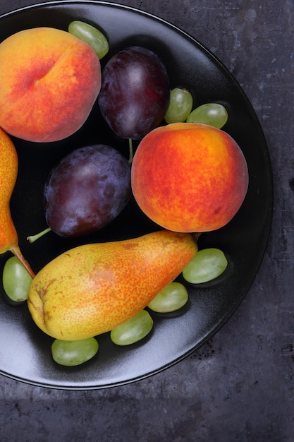 Group of healthy summer colorful fruits on a dark background closeup Pears peaches plums and grapes on a black plate View from above