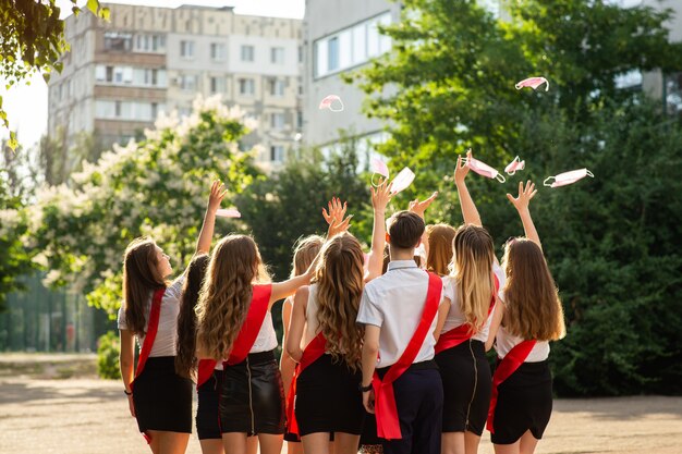 group of high school graduates in smart school uniforms and red ribbons happily throw up medical masks as a symbol of the end of quarantine.