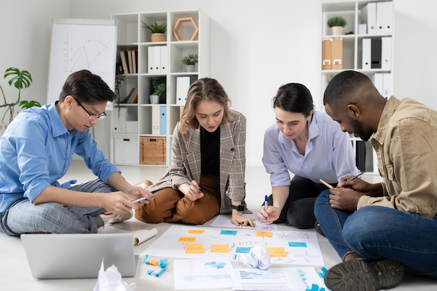 Group of interracial startuppers sitting on floor with task board and analyzing marketing strategies in office