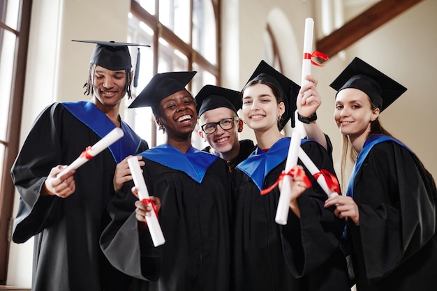 Group of joyful young people wearing graduation gowns at\
ceremony in university and smiling at camera