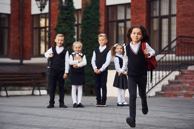 Group of kids in school uniform posing to the camera outdoors together near education building.