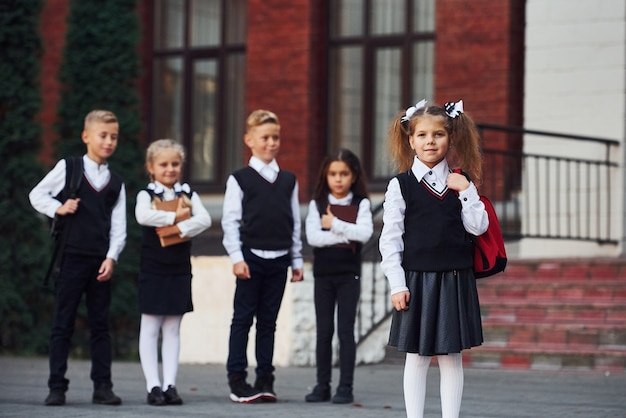 Group of kids in school uniform posing to the camera outdoors together near education building.