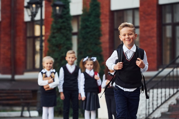 Group of kids in school uniform posing to the camera outdoors together near education building.