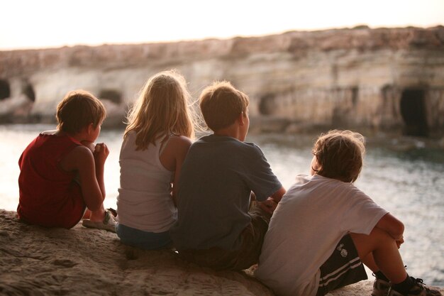 a group of kids sit on a rock and look at the water