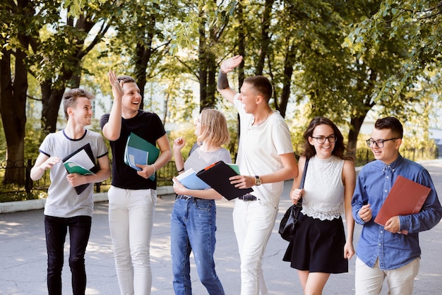 Group of laughing students with books outdoors on a bright sunny day park and building of university