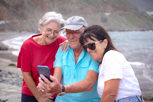 Group of mature friends sitting at the beach enjoying the day outdoors while they all look at a cellphone Seniors people enjoying retirement free time and vacation