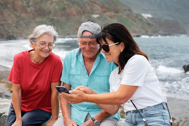 Group of mature friends sitting with their backs to the sea enjoying the day outdoors while they all look at a cellphone Seniors people enjoying retirement and free time