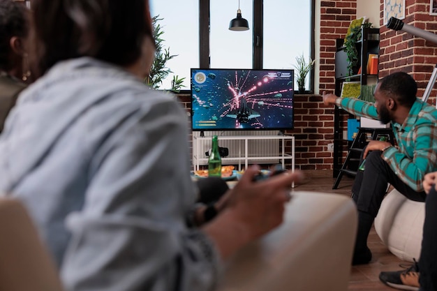 Group of men and women having fun with shooter video games on tv, using console to play on television. Playing shooting game at social gathering with friends, enjoying leisure activity and gaming.