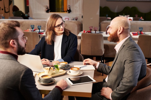 Group of middle-aged managers sitting at table with coffee and discussing ideas during business meeting in cafe