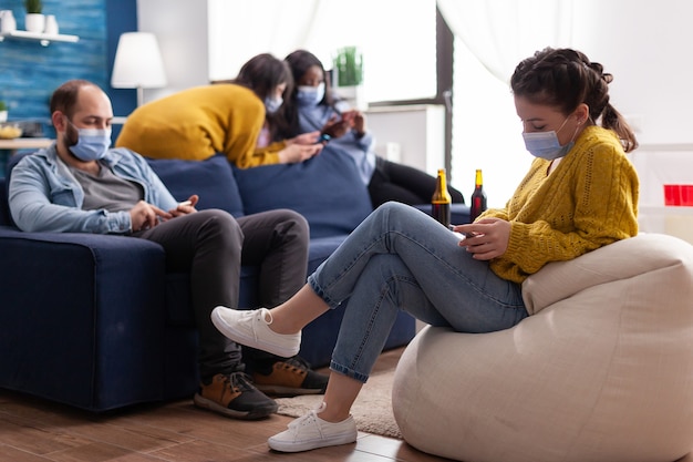 Photo group of mixed race friends browsing on smartphone keeping social distancing wearing face mask to prevent spread of coronavirus during global pandemic in living room with beer bottle. conceptual image