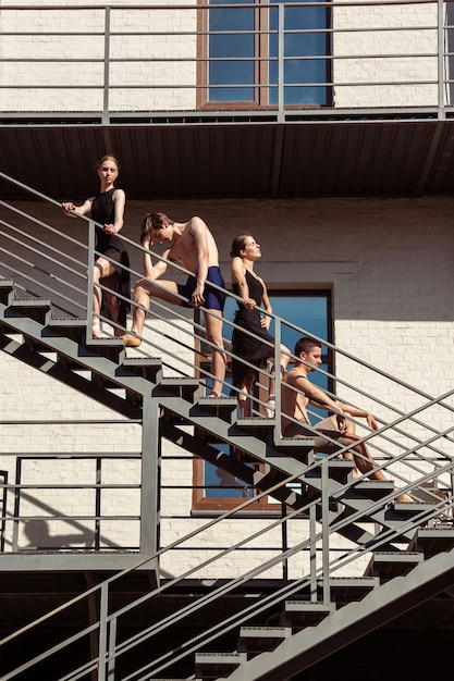 The group of modern ballet dancers performing on the stairs at the city. Fast moving of citylife, grace in daily things around. Graceful and flexible models in sunshine with buildings on background.