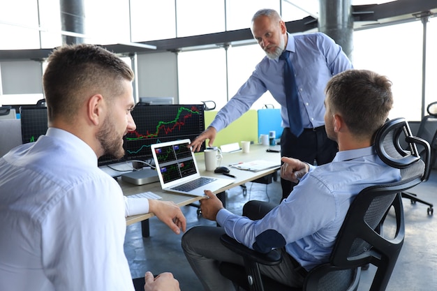 Gruppo di uomini d'affari moderni in abiti da cerimonia che analizzano i dati del mercato azionario mentre lavorano in ufficio.