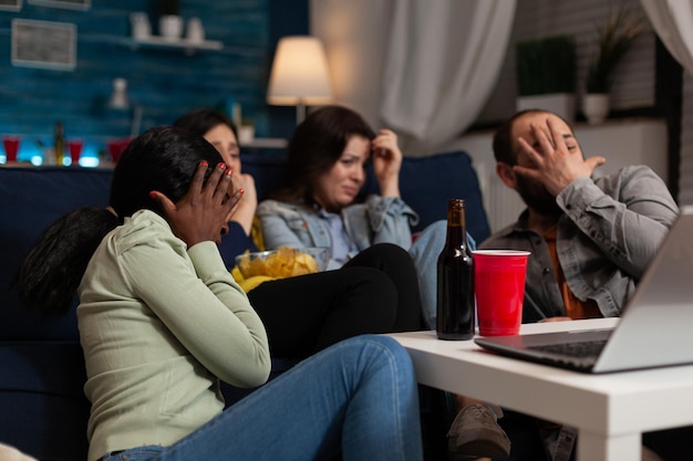 Group of multi-ethnic friends watching horror thriller movie on\
laptop computer making scared facial expresion during film night\
sitting on couch in living room. hanging out concept