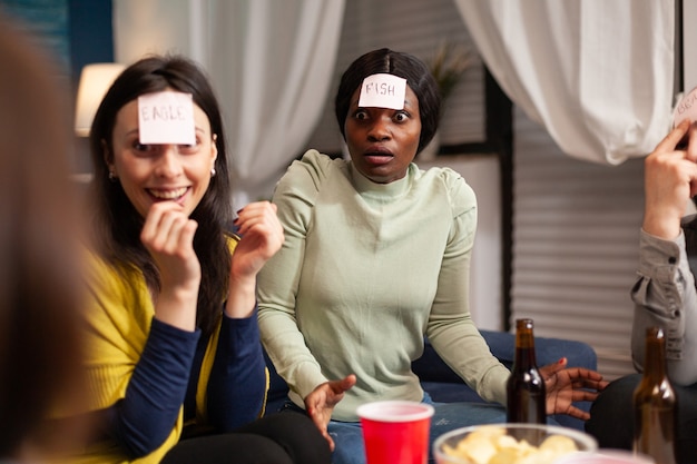 Gruppo di persone multietniche che si divertono mentre giocano a giochi da tavolo usando note adesive che trascorrono del tempo insieme. amici creativi che escono a tarda notte in soggiorno durante la festa in casa.