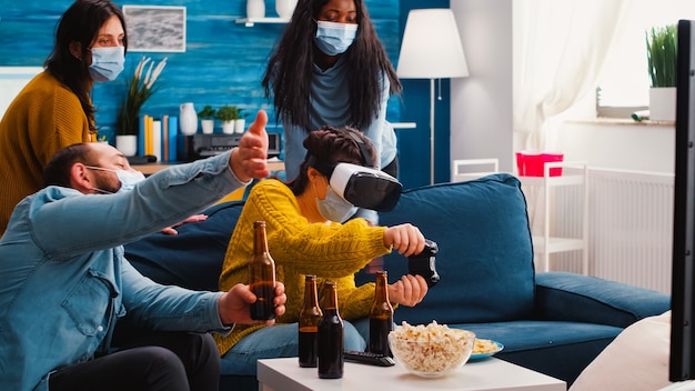 Group of multiethnic friends having fun playing video game
experiencing virtual reality headset keeping social distancing in
home living room due to social pandemic