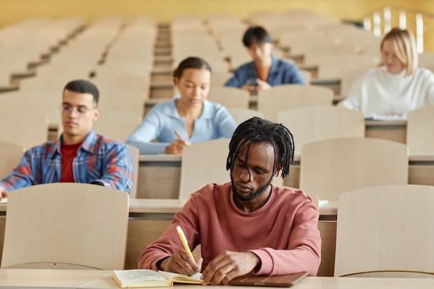 group of multiethnic students making notes in notebooks while sitting at desk during lecture