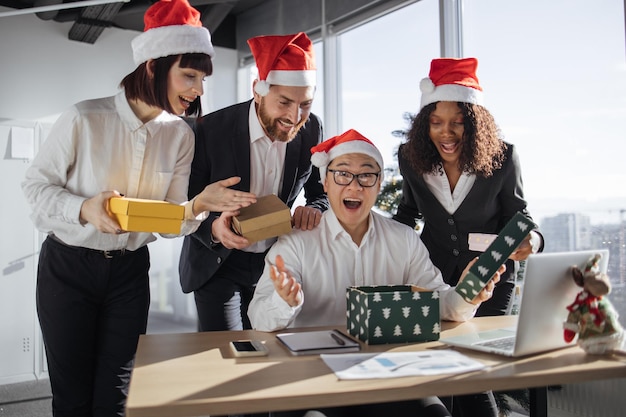 Photo group of multinational business colleagues in santa hats giving christmas gift boxes to asian male manager sitting at table unboxing present and looking with surprise at camera