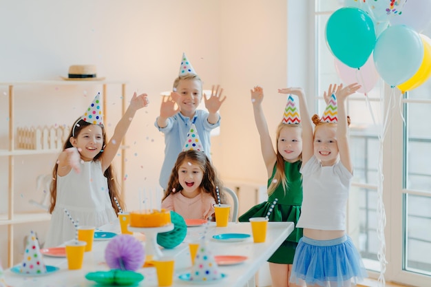사진 행복한 아이들의 그룹은 풍선이 있는 장식된 방에서 축제 테이블 근처에서 색종이 조각을 하고 파티 모자 포즈를 취하며 즐거운 표정을 짓고 함께 생일을 축하합니다.
