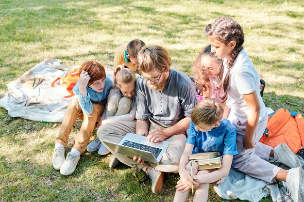 Фото Группа улыбающихся маленьких детей, обучающихся на открытом воздухе, сидя в ряду на зеленой траве и писать в учебниках