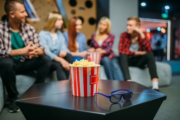 Группа подростков отдыхает на диване и ждет сеанса в кинозале. мужская и женская молодежь, сидя на диване в кинотеатре, попкорн на столе