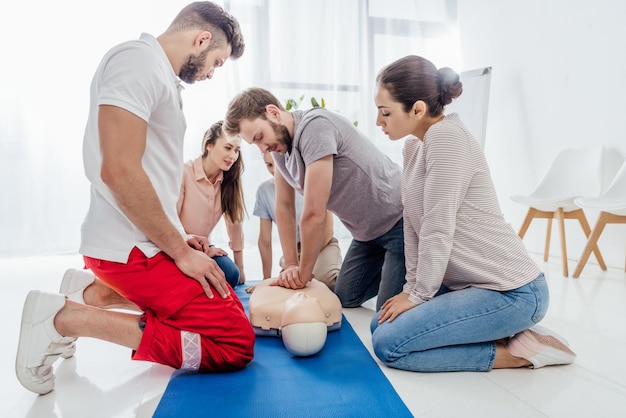 Группа людей смотрит на мужчину, выполняющего сердечно-легочную реанимацию на манекене во время обучения оказанию первой помощи