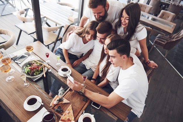 A group of people make a selfie photo in a cafe. The best friends gathered together at a dinner table eating pizza and singing various drinks.