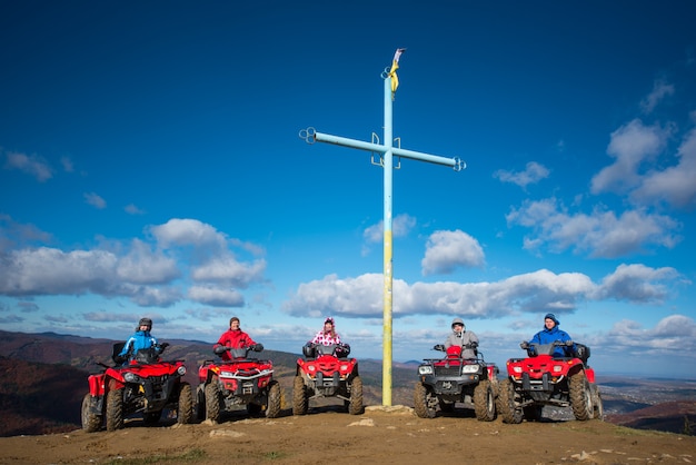 Group of people on red quad bikes near cross with a symbol of Ukraine on top of the mountain 