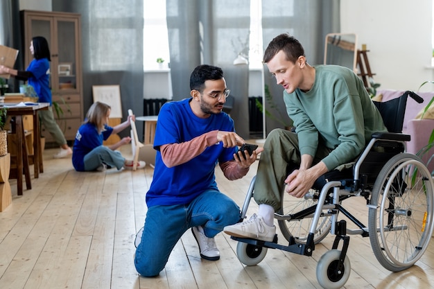 A group of people sitting in a room while young male volunteer sitting on squats in front of guy in wheelchair