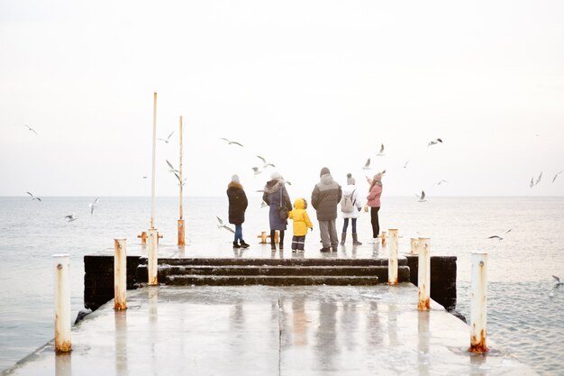 A group of people in winter clothes are standing on the dock and feeding the gulls from their hands.