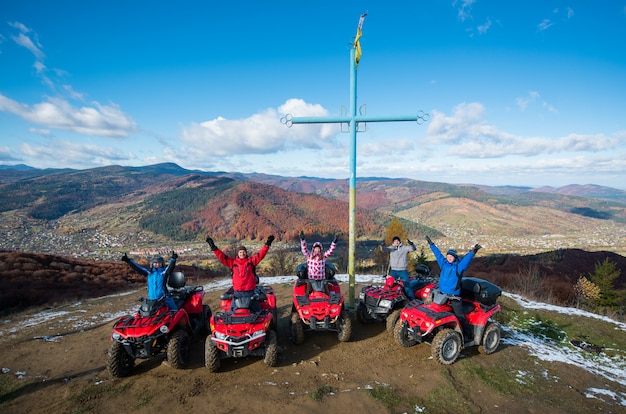Группа людей с поднятыми руками вверх на красных квадроциклах возле креста с символом Украины на вершине горы