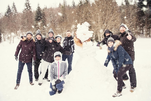 group portrait of young happy business people after a competition posing with finished snowman while enjoying snowy winter day with snowflakes around them during a team building in the mountain forest