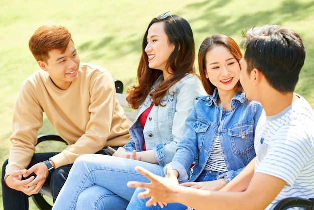 Group of positive university students sitting outdoors on campus and discussing new classes and vaca...