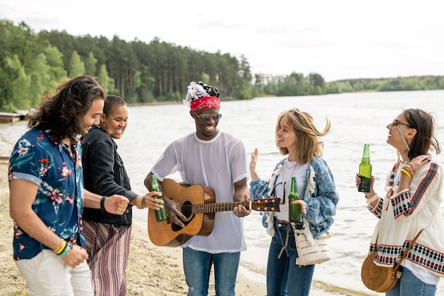 ヒッピー黒人の男によるギター音楽の下でビーチで踊るポジティブな若い多民族の友人のグループ