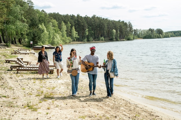 Gruppo di giovani amici multietnici positivi che godono della passeggiata sulla spiaggia mentre il ragazzo nero suona la chitarra in movimento